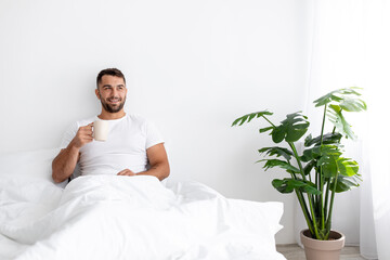 Happy glad millennial caucasian handsome muscular male sits on white bed, drinks coffee, enjoy good...