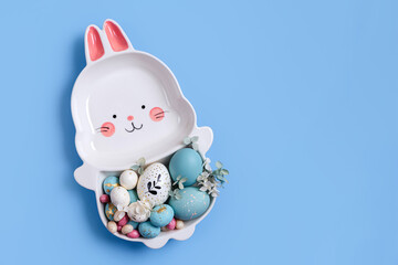 Cute plate in the shape of a bunny with  Easter eggs and candy on blue background. Happy Easter concept.