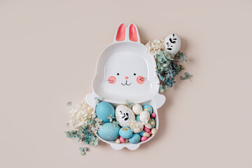 Cute plate in the shape of a bunny with  Easter eggs, candy and flowers on beige background. Happy Easter concept.