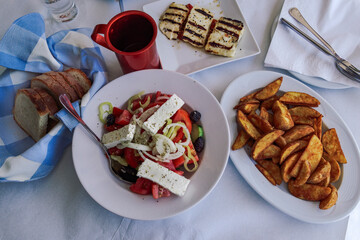 Close up of Horiatiki Greek salad as served on a tavern. Top view of restaurant table with with feta cheese, tomato, cucumber, onions, olives, wine, bread and fries.