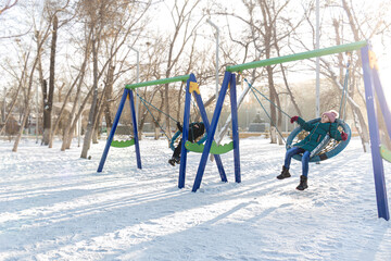 children boy and girl ride on a swing in the park in winter.
 winter walk in the park.lifestyle, winter clothes 

