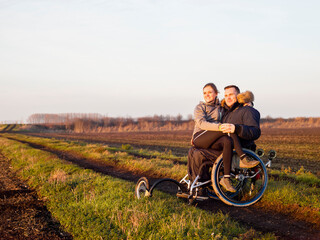 Smiling man on wheelchair in field with woman on laps - obrazy, fototapety, plakaty