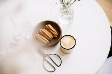 Delicious fresh cappuccino coffee with three eclairs and scissors on the white table background and some gypsophila in a vase, top view, flat lay