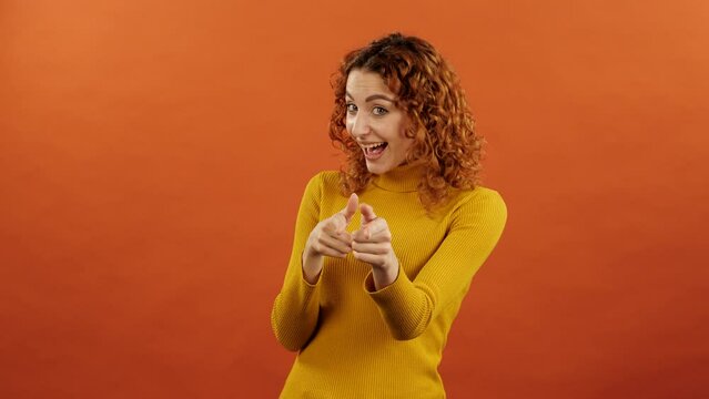 Attractive redhead caucasian girl in orange jumper pointing fingers at you and laughing isolated over orange studio background.