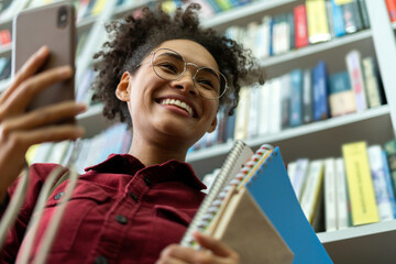 Young and beautiful businesswoman holding a lot of books in her hands and standing near the bookshelves in the modern office and using smartphone with wide smile