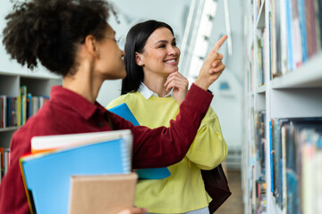 People, knowledge, education and school concept. Happy student girl or young woman taking book from shelf in library while standing near her classmate