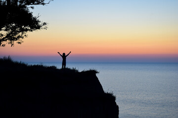 Silhouette of an unrecognizable woman with outstretched arms near the branches of a tree against the backdrop of the evening sea