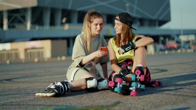 At sunlight two young women sitting on ground with roller skates use phone smiling feel happy street in summertime legs rollerblading activity slow motion