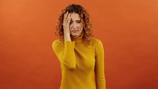 Attractive caucasian redhead girl in an orange jumper slapping her face with her hand because of a mistake and bankruptcy isolated on orange studio background.