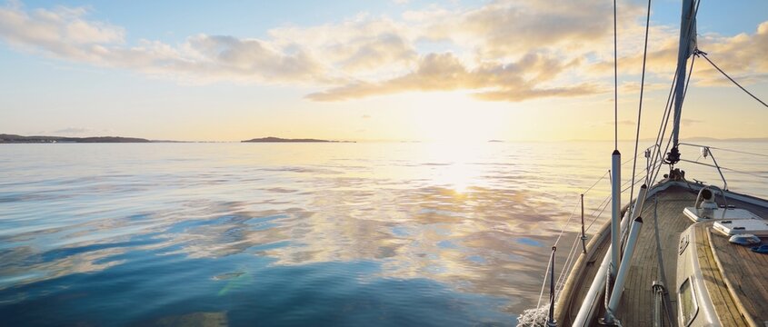 Sloop rigged modern yacht with wooden teak deck sailing at sunrise. A view from the deck to the bow. Colorful clouds. Port Ellen, Isle of Islay, Inner Hebrides, Scotland, UK