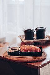 Delivered sushi with chopsticks and plastic box with salmon roll with two cups of coffee on a table. Cozy home. Eating at the kitchen. Meal. Traditional Asian food. Takeout and delivery.