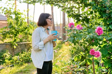 Middle-aged happy woman relaxing in garden with a cup in her hands, enjoying blooming rose