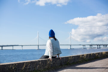 A hipster girl in a blue hat and a blue vest on the shore of a bay or sea. The girl looks into the distance at the water and a beautiful bridge