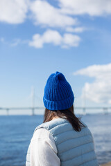 A hipster girl in a blue hat and a blue vest on the shore of a bay or sea. The girl looks into the distance at the water and a beautiful bridge