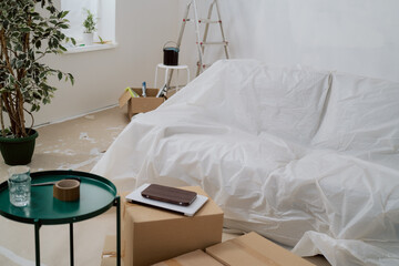 Interior of the room during renovation painting walls with ladder, floor and couch protected with foil cardboard, moving