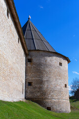 Round fortress tower with loopholes at the junction with the fortress wall. A tower with a hipped wooden roof. Side view. Early spring. Daylight. Clear sky.