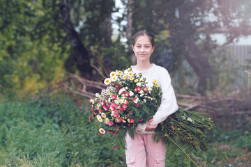 A girl holds in her hands a large beautiful bouquet of autumn flowers in a white sweater in a forest glade.