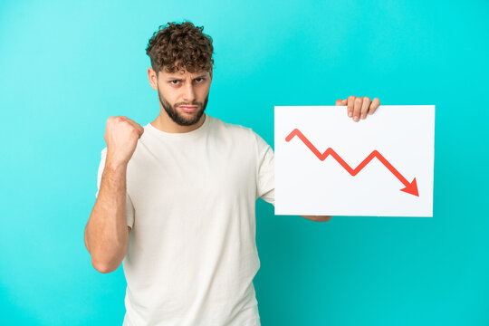 Young handsome caucasian man isolated on blue background holding a sign with a decreasing statistics arrow symbol and angry