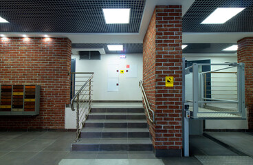 The hall of a modern multi-storey building with stairs and a lift for the disabled. Brick finish. Mailboxes. Grey tile.