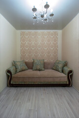 A green-beige classic sofa with pillows in a room with a chandelier. The sofa is surrounded on three sides by walls. Wallpaper in moldings. Stretch ceiling. Laminate flooring on the floor.