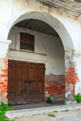 View from the corner of the arch of the old building of the shopping mall. Closed door. Boarded up window. White peeling walls. A red brick is visible. Summer. Daylight.