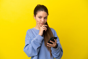 Little girl over isolated yellow background using mobile phone and thinking