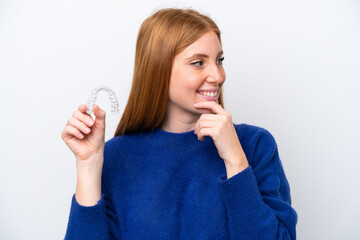 Young redhead woman holding invisible braces isolated on white background thinking an idea and...