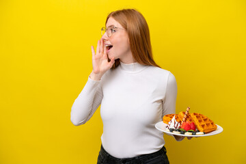 Young redhead woman holding waffles isolated on yellow background shouting with mouth wide open to the side