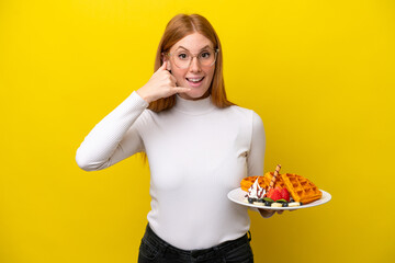 Young redhead woman holding waffles isolated on yellow background making phone gesture. Call me back sign