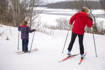 Mother and young daughter on cross-country skis at the top of a hill, learning to ski, daytime, cloudy, snowy river in background