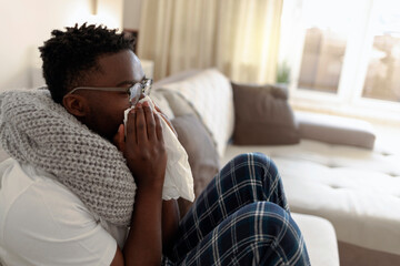 Young man sitting in his bed and blowing his nose while suffering from a cold. Sick African with cold or flu virus is sitting under the blanket on the sofa and blowing his nose with paper tissues.