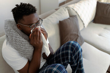 Sick man lying on sofa at home in living room. Young African man feeling sick with cold and fever...