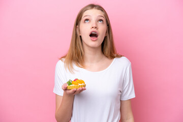 Teenager Russian girl holding a tartlet isolated on pink background looking up and with surprised expression