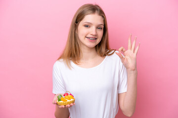 Teenager Russian girl holding a tartlet isolated on pink background showing ok sign with fingers