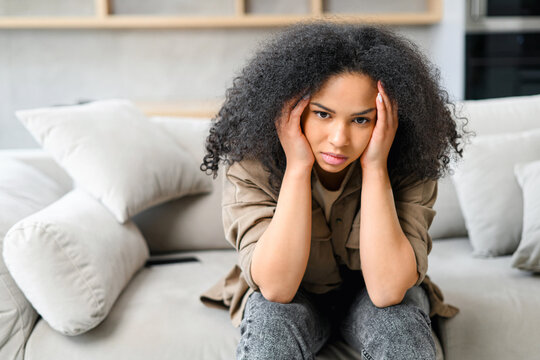 Frustrated african-american woman sitting on the sofa in grief, disappointed multiracial female feels despair, worrying, depression, loneliness, suffering from strong headache