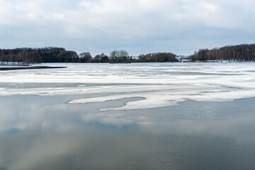 Beautiful winter landscape forest on the lake shore on a sunny frosty day. Panorama of the coastline covered with snow and birch grove. Ice on the river and the reflection of clouds in the water.