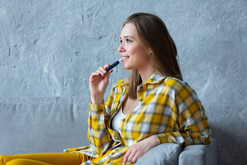 Profile photo of a smiling beautiful girl with an electronic cigarette in her mouth resting on a blue sofa at home