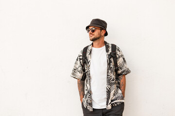 Hipster man in fashion summer clothes with backpack stands near white wall. Guy travel on the street