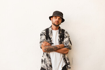 hipster stylish man with a bucket hat in fashion beachwear with a shirt and a backpack stands near...