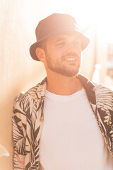 Happy handsome hipster man with smile in fashion shirt with palm trees print and black bucket hat at sunset