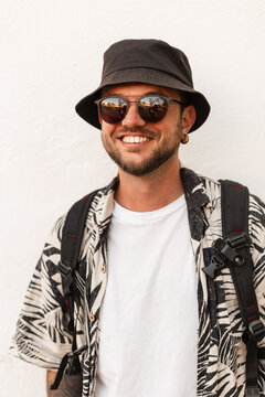 Happy smiling hipster man with smile in fashion summer clothes with sunglasses and hat stands near white wall