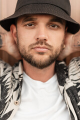 Close-up street portrait of handsome young hipster man with beard in fashion beachwear clothes with bucket hat stands near wall