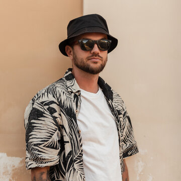 Handsome stylish hipster man with beard, sunglasses and bucket hat in fashion  summer outwear with beach shirt near the wall on the street Stock Photo