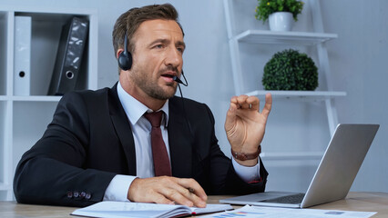 man in headset talking and gesturing near laptop at workplace