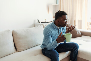 Unhappy handsome young businessman sitting alone in his home office and talking on his cellphone. African young man in eyeglasses sitting on couch, drinking coffe while using his cellphone at home.