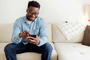 Handsome young African man looking on phone and smiling while sitting on the couch at home. Young happy black man at home sending messages on smartphone lying on beige couch in living room, copy space