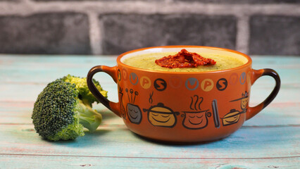 on a blue wooden table there is a brown plate with green broccoli puree soup with dried tomatoes on...