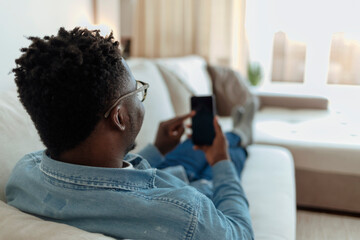 Handsome young African man looking on phone and smiling while sitting on the couch at home. Young...