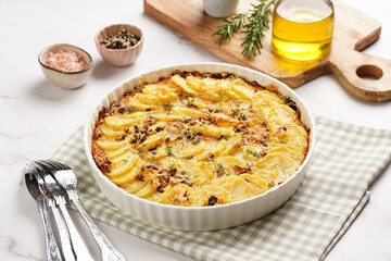 Slices potato casserole with red onions, fresh thyme and rosemary on green checkered kitchen towel , wooden board with flavoring ingredients: pepper, salt, olive oil