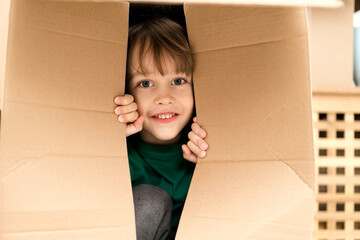 Boy hiding in inside a huge cardboard box. He is playing and peeking through a hole in box. Kid is happy about moving into a new home.